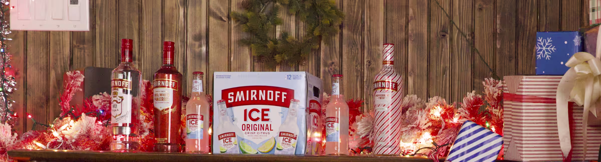 Festive display with Smirnoff Ice and vodka bottles, surrounded by holiday lights, wreath, and wrapped gifts against a wooden backdrop.