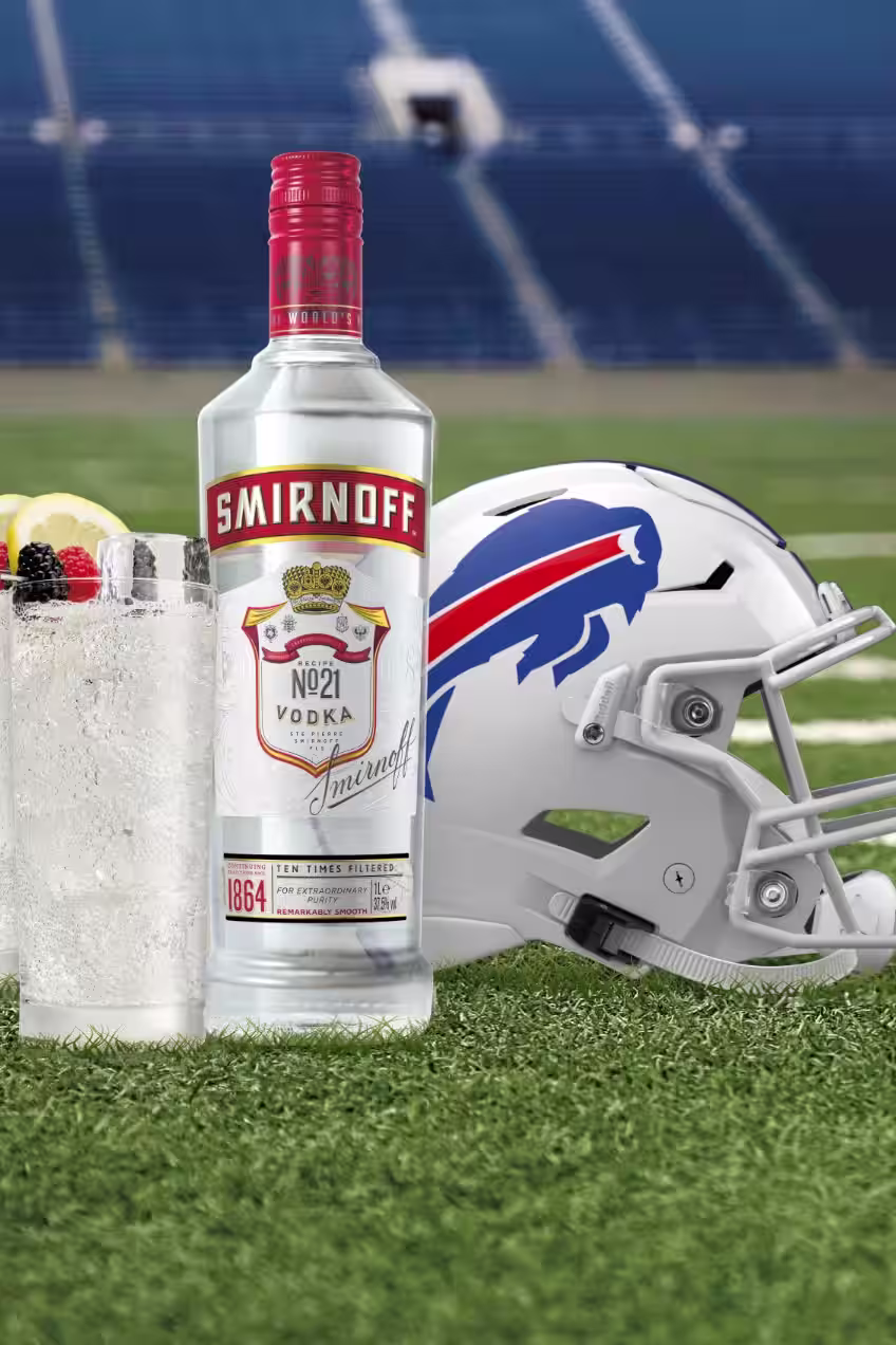A bottle of Smirnoff No. 21 Vodka and two Smirnoff Red, White & Bills cocktails garnished with lemon slices and berries are placed on a football field next to a Buffalo Bills helmet.