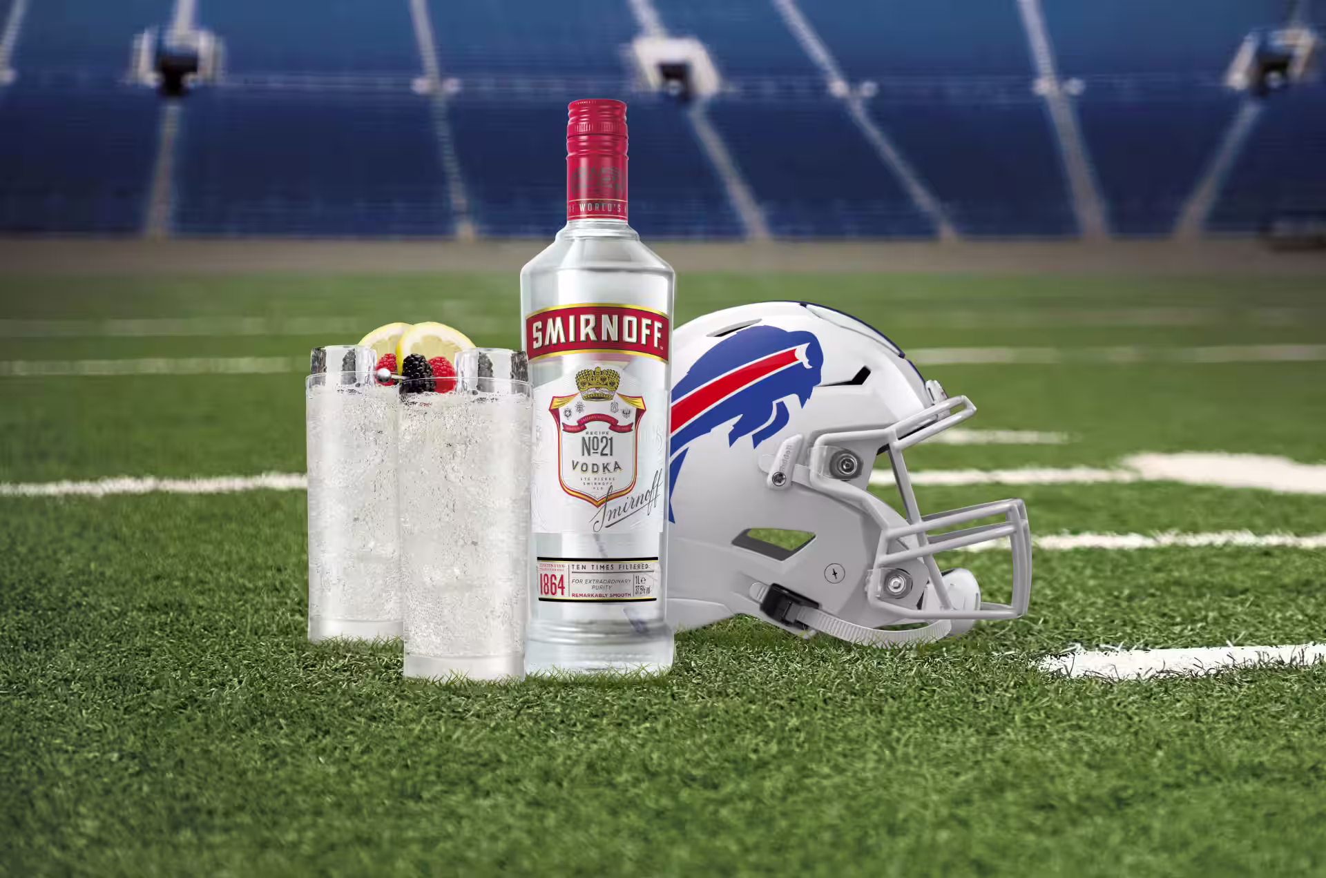 A bottle of Smirnoff No. 21 Vodka and two Smirnoff Red, White & Bills cocktails garnished with lemon slices and berries are placed on a football field next to a Buffalo Bills helmet.