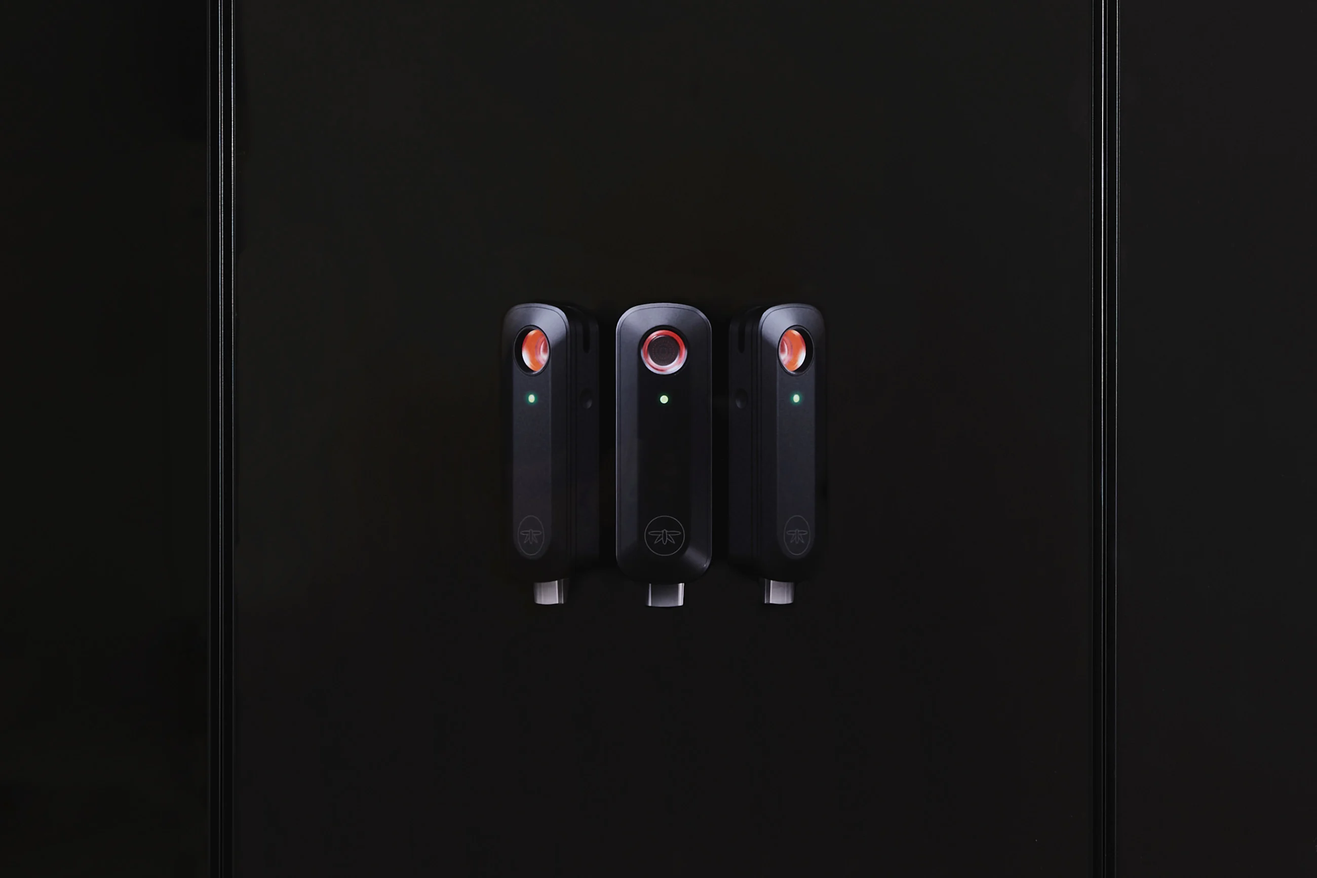 Three black electric vehicle chargers are mounted on a dark wall, each with a small green indicator light.