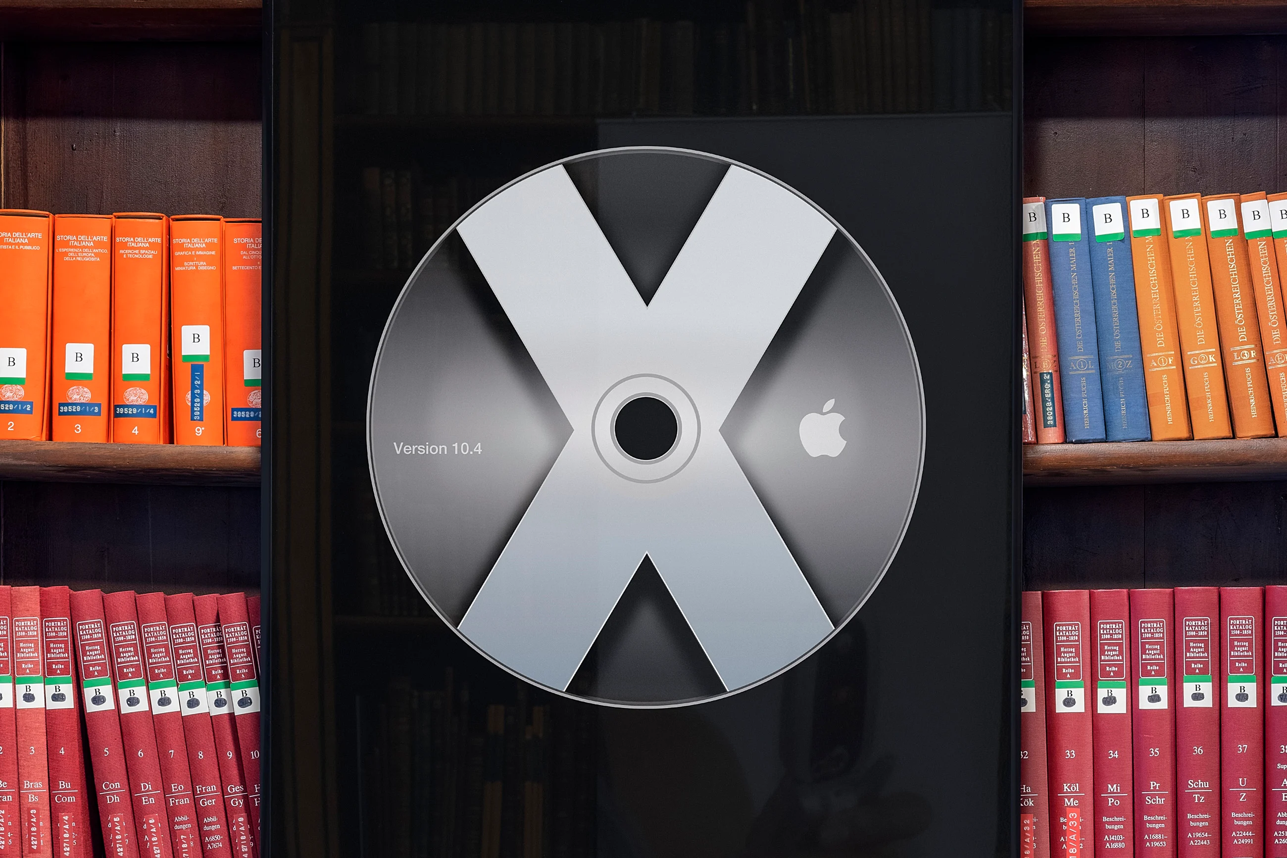 A large CD with a bold X and Apple logo is displayed against a background of shelves filled with orange and red books.