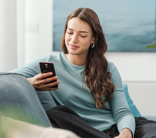 A picture of a woman sitting on a sofa and looking at her phone. The woman is smiling.