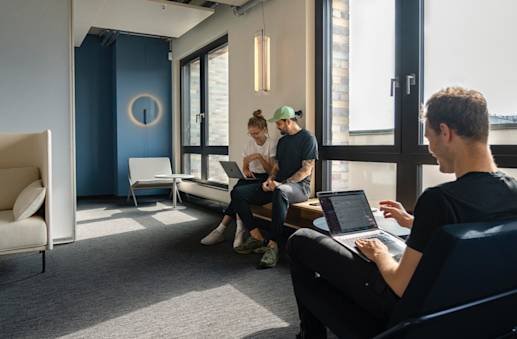 Eine Büroumgebung, in der ein Mann sitzt und an seinem Notebook arbeitet, während im Hintergrund eine Frau und ein Mann ein Projekt besprechen und dabei in ein Notebook schauen.