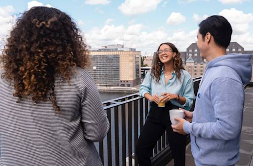 Drei Personen - zwei Frauen und ein Mann - unterhalten sich auf einem Balkon an der Spree in Berlin. Eine Frau lächelt.