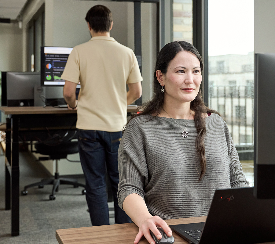Picture of woman in the workplace sitting in front of a desktop. There's a man working in the background.
