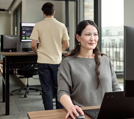 Bild von einer Frau am Arbeitsplatz, die vor einem Schreibtisch sitzt. Im Hintergrund arbeitet ein Mann.