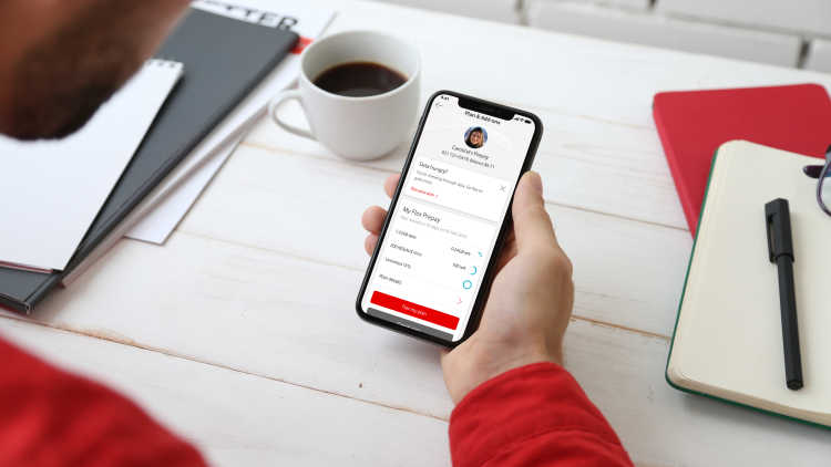 A bearded man in a long-sleeved red shirt sits at a table, holding his phone with a red-themed app open on the screen.