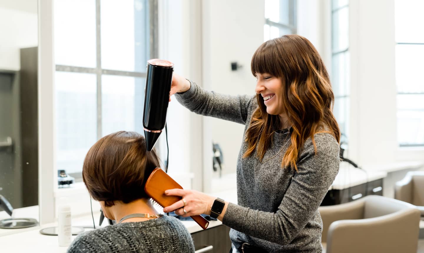 A smiling hairdresser blowdries and brushes the hair of a seated client inside a hair salon.