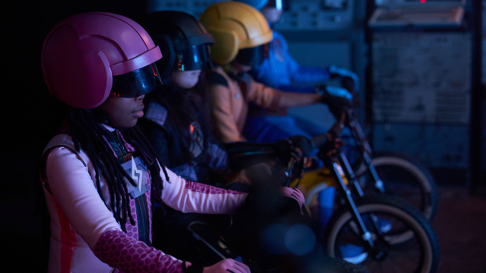 Three kids wearing matching helmets and suits in pink, black and yellow lined up on the Youth Series bike getting ready to ride