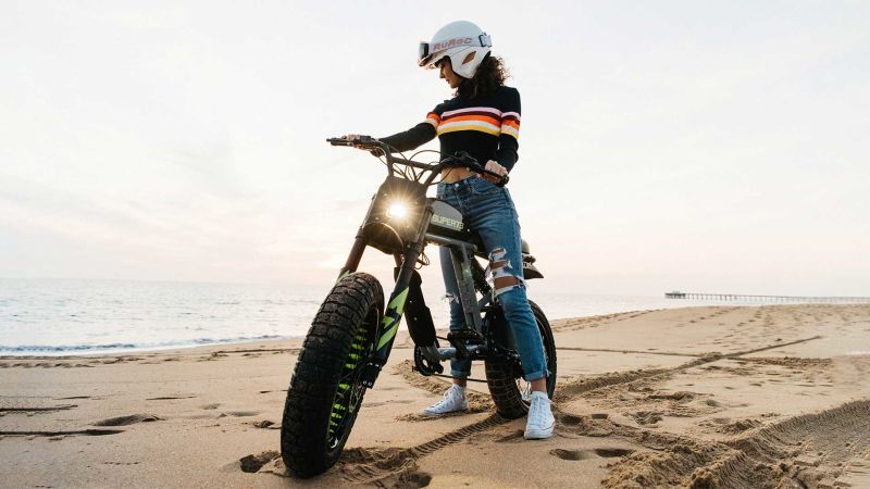 A young woman riding a Super73-RX on the beach with the headlight shining forward.