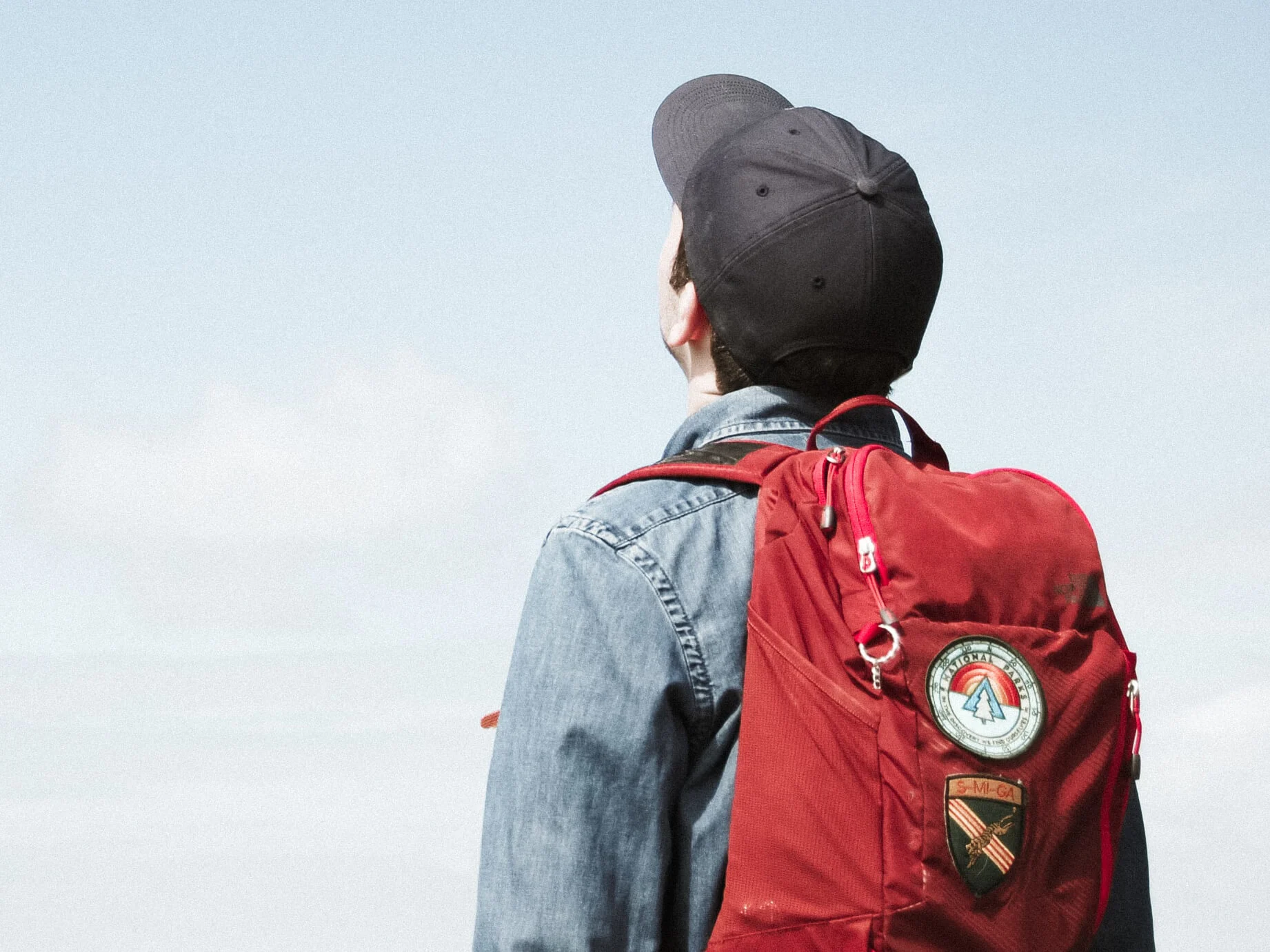 A young person wearing a red backpack and black baseball cap looks up at the sky
