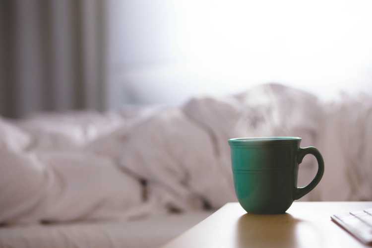 Green coffee mug on side table with a messy bed in the background.