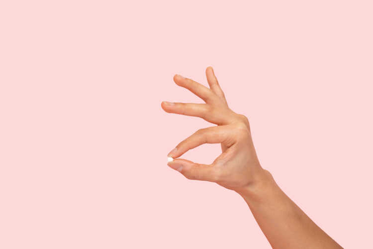 Hand holding a Plan B pill between thumb and forefinger on pink background.