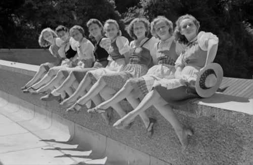 A black and white photo of a line of women sitting on a ledge, kicking out their legs.