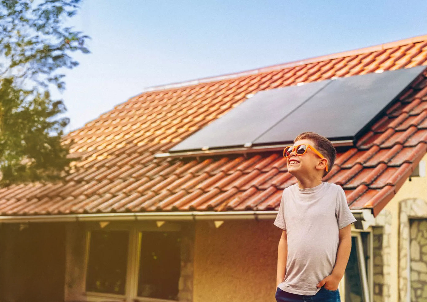 Kind mit Sonnenbrille lacht vor einem Haus mit Photovoltaik in den Himmel