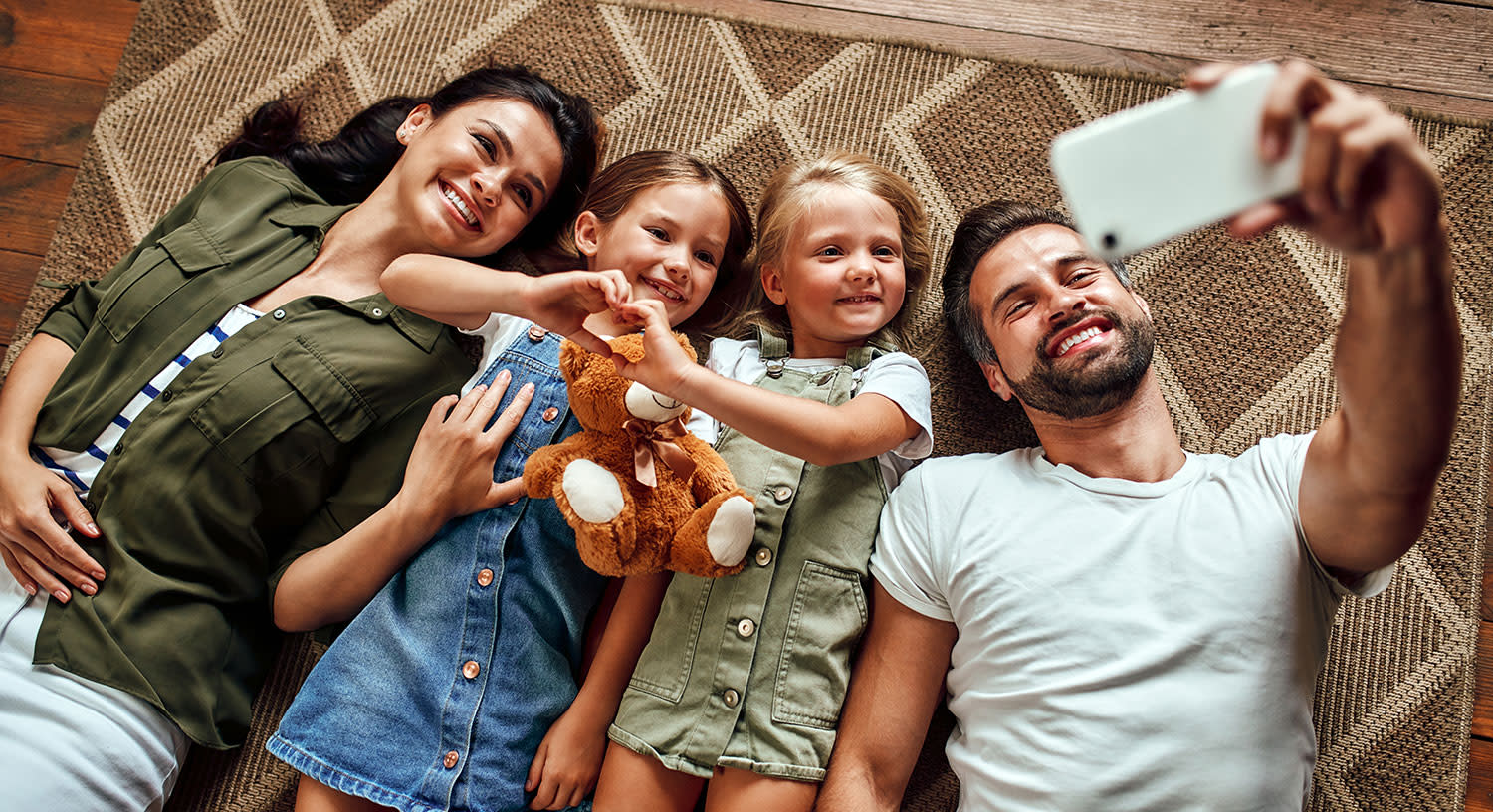 Vater, Mutter und 2 Töchter liegen auf einem Teppich und schauen auf ein Smartphone