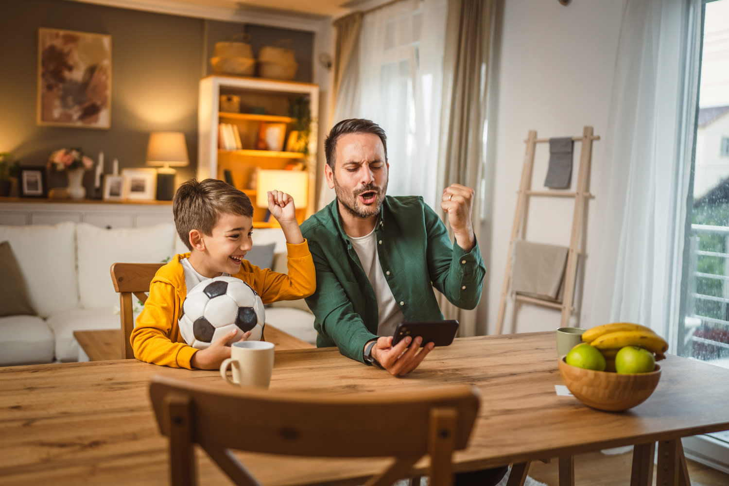 Ein Mann und sein Sohn sitzen im Esszimmer am Tisch und schauen voller Freude auf dem Handy ein Fußbalspiel an