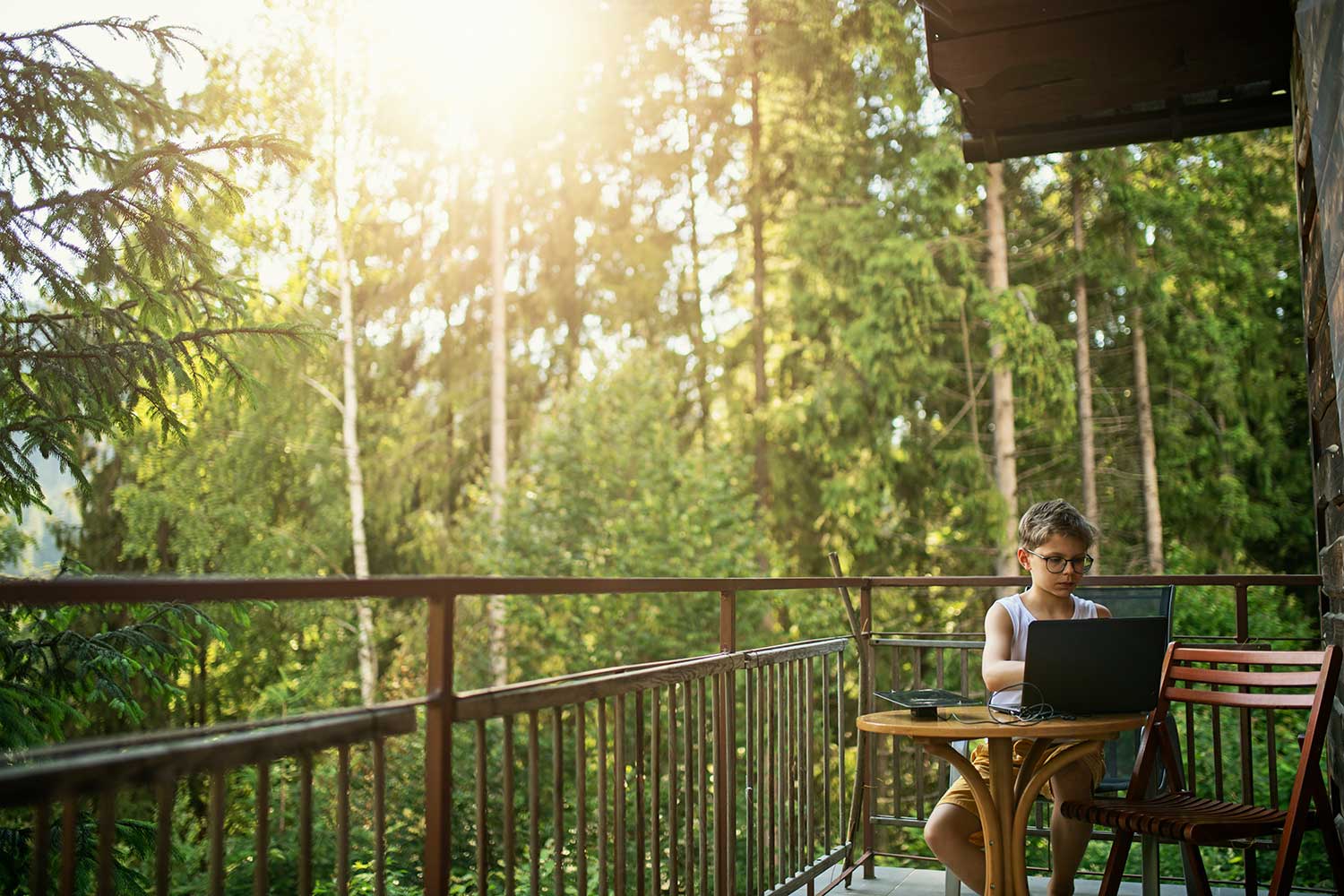 Kleiner Junge sitzt mit Laptop auf dem Balkon mit Wald im Hintergrund