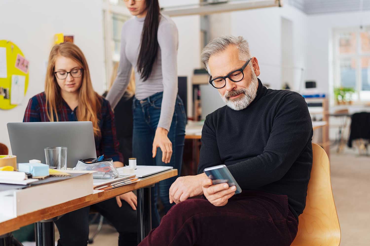 Mann sitzt am Handy im Büro, Kolleginnen arbeiten am Laptop im Hintergrund.
