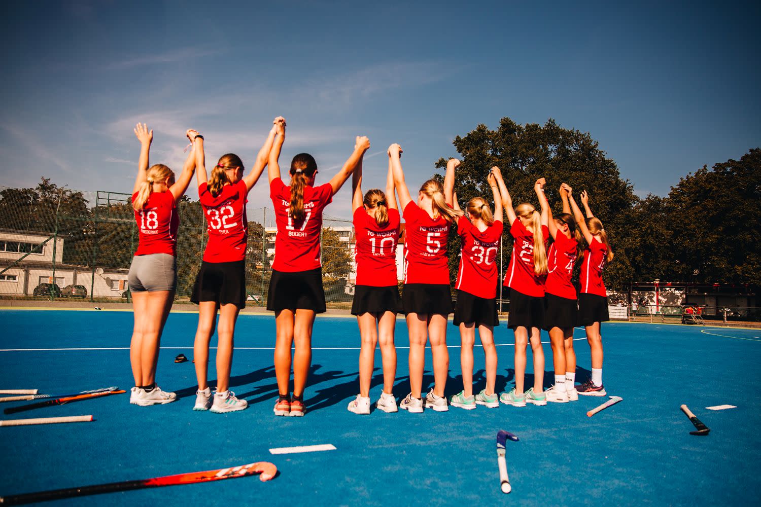 Eine Mädchengruppe steht auf einem Hockeyplatz im Halbkreis und halten die Hände hoch Eine Mädchengruppe steht auf einem Hockeyplatz im Halbkreis und halten die Hände hoch