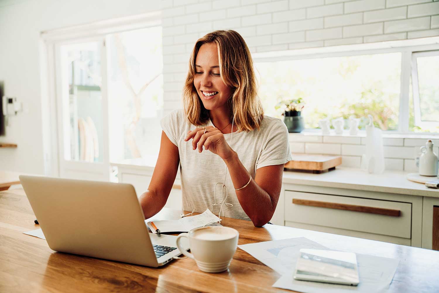 Frau sitzt mit Ihrem Laptop und einer Tasse Kaffee in der Küche.