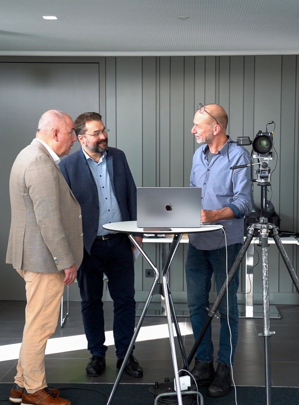 Drei Personen stehen an einem Stehtisch und besprechen sich vor einem Laptop, neben ihnen ist eine professionelle Kamera auf einem Stativ aufgebaut