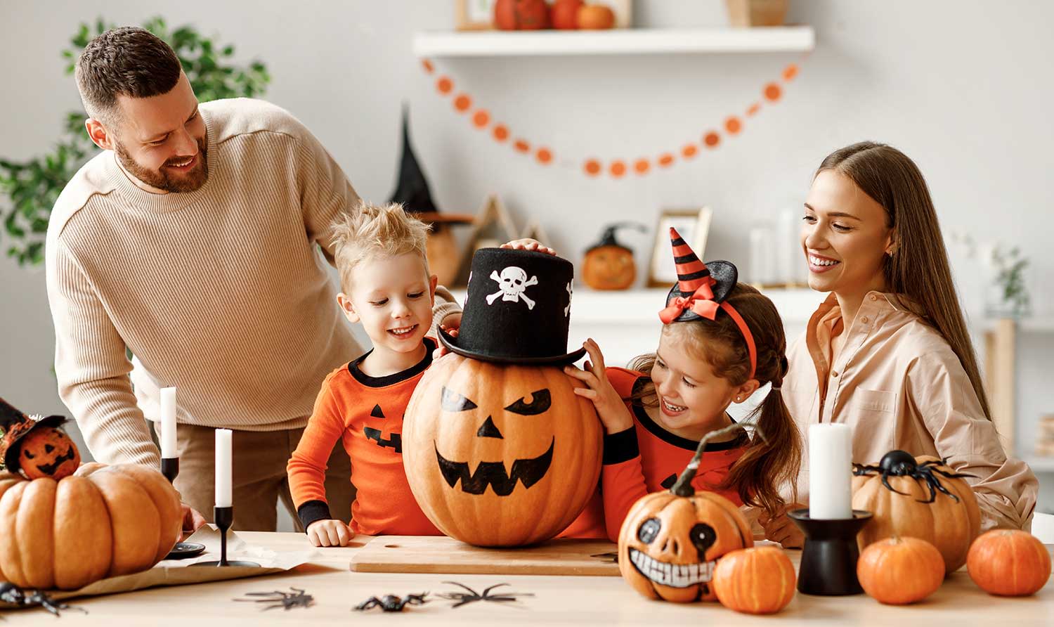Titelbild Eine Familie sitzt an einem Tisch und bereitet sich auf Halloween vor. Bemalte Kürbisse sind auf dem Tisch platziert