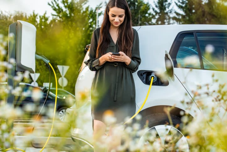 Frau mit Handy, während sie ihr Elektroauto auf dem Parkplatz auflädt