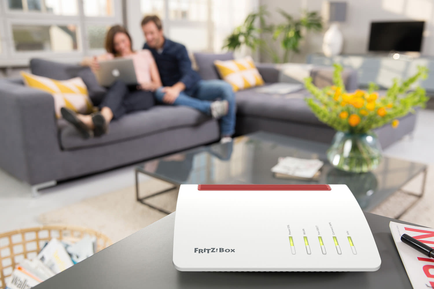 Router AVM-FRITZ-Box-7590 steht auf einem Tisch. Personen sitzen im Hintergrund auf dem Sofa