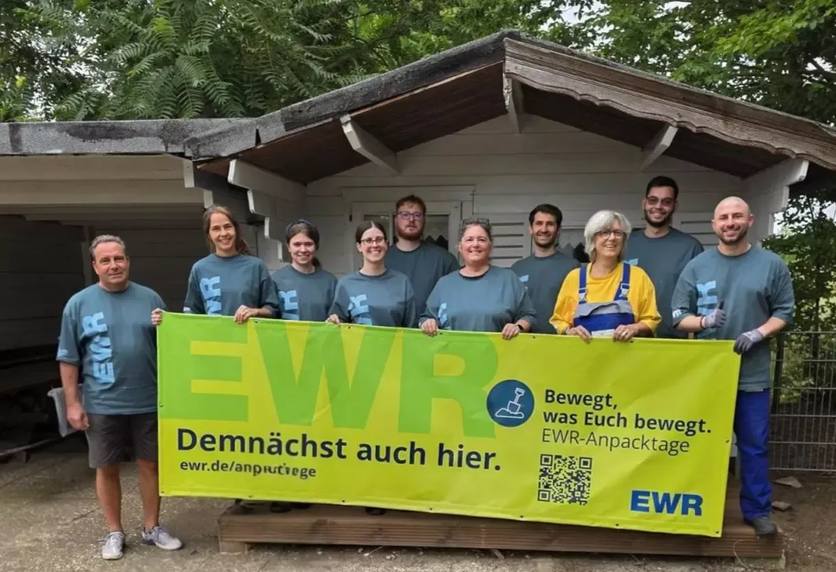 EWR Mitarbeiter halten Schild vor renovierter Holzhütte.