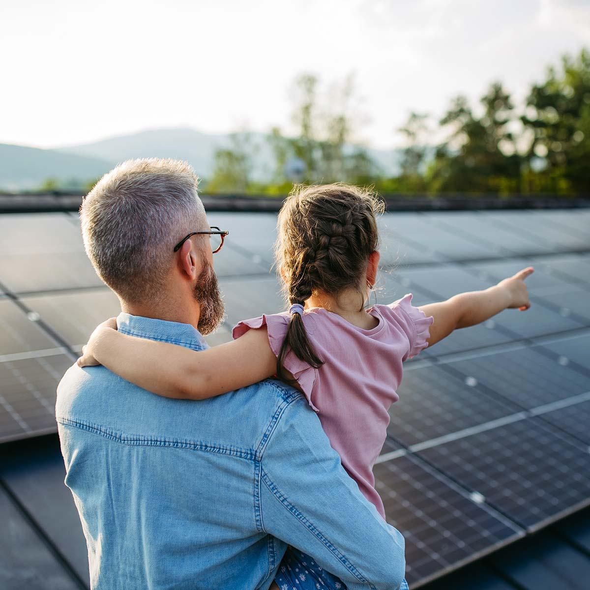 Ein Mann mit Kind auf Arm betrachtet Photovoltaikanlage auf Hausdach
