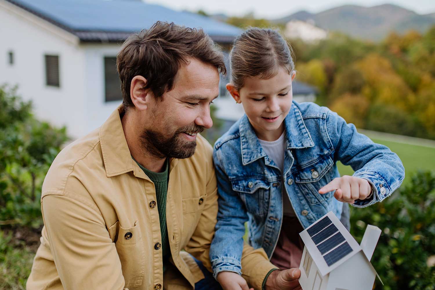 Mann und Tochter stehen an Papierhaus mit Photovoltaikanlage im Garten