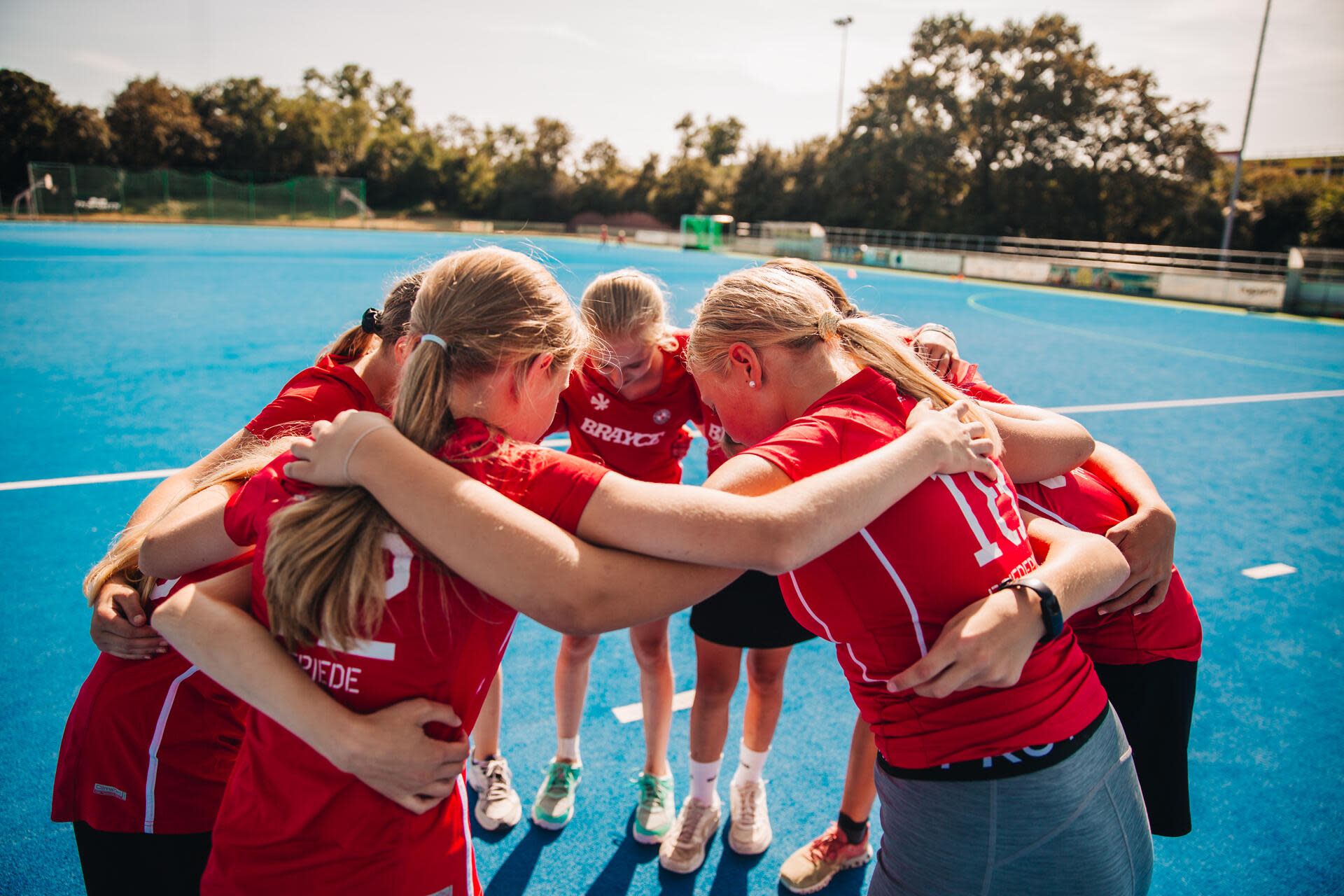 Ein Hockeyteam steht eng im Kreis auf einem blauen Hockeyfeld, alle legen die Arme umeinander. Ein Hockeyteam steht eng im Kreis auf einem blauen Hockeyfeld, alle legen die Arme umeinander.