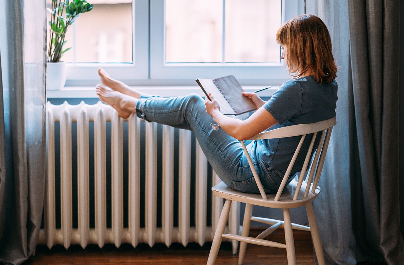 Frau sitzt barfuß an einer Heizung und liest ein Buch