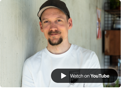 A photo of the SumUpper Ben smiling with a short beard, wearing a white t-shirt and black cap, leaning against a light concrete wall. A 'Watch on YouTube' button overlays the image, indicating a video feature
