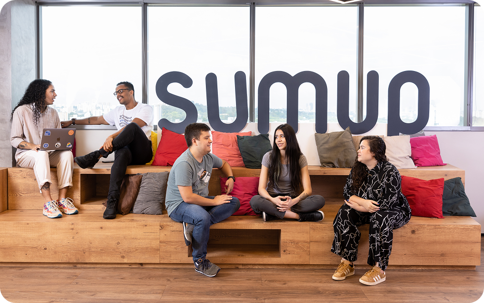 SumUp employees in a vibrant South American office chatting on tiered wooden seating in front of a large SumUp logo. The diverse team sits casually with laptops and pillows, highlighting a culture of collaboration, inclusivity, and creativity.