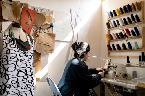 Person using a sewing machine in a well-lit room. Spools of thread line the wall, and sewing patterns hang nearby. A dress form with a patterned garment stands on the left.