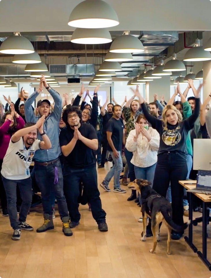 A lively group of SumUp employees in an open office claps and cheers while posing playfully. Overhead lights brighten the space, and a dog walks in the foreground, reflecting SumUp’s fun and inclusive workplace culture in Santiago, Chile.