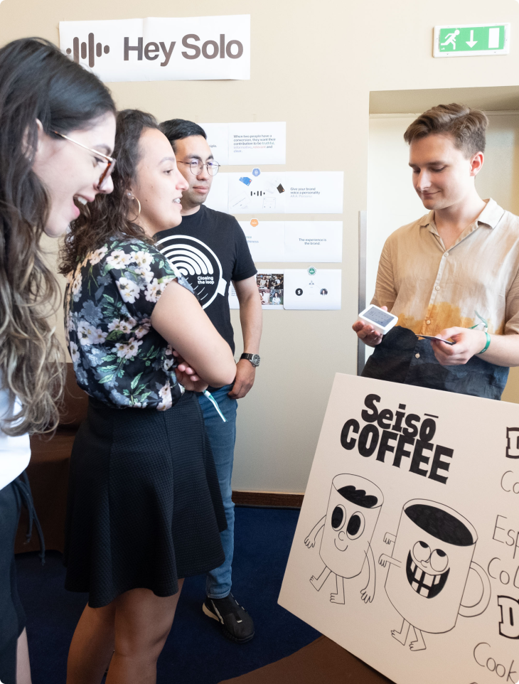 Four SumUp team members interacting during Hack Week, with one person demonstrating a project called 'Seisō Coffee' displayed on a poster featuring illustrated coffee mugs, while another project display labeled 'Hey Solo' is visible in the background.