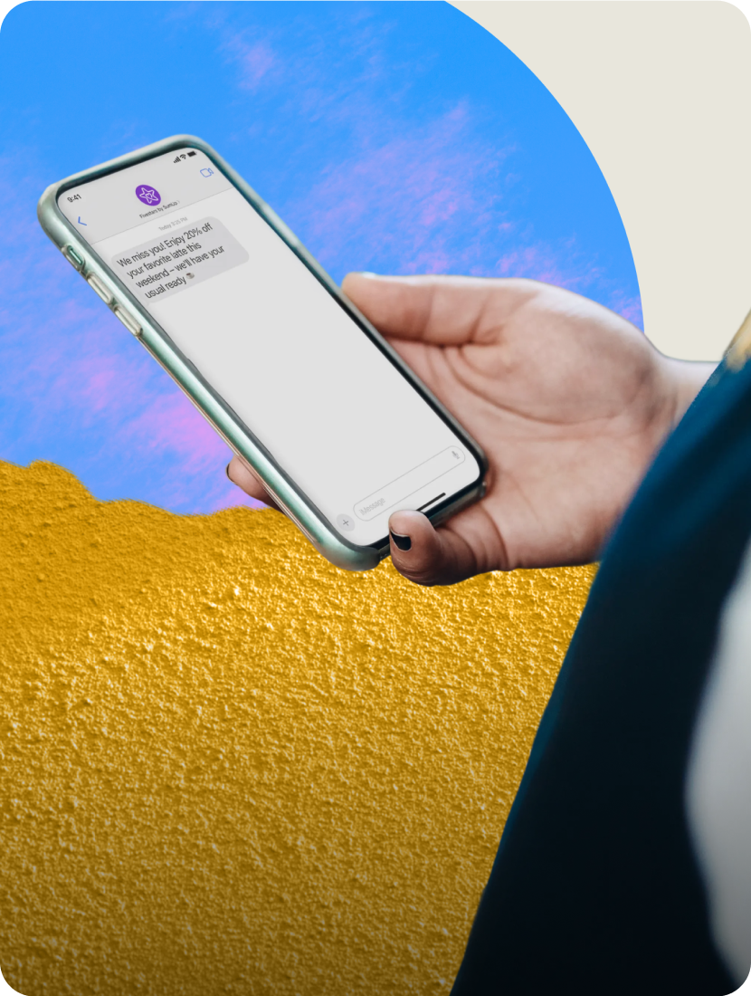 A hand holds a smartphone displaying a text message: "We miss you! Enjoy 20% off your favorite latte this weekend โ We have your usual ready." The background appears blurred.