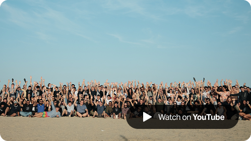 Large group of SumUp team members on a sandy beach with their arms raised in celebration during Hack Week 2023, with a 'Watch on YouTube' button overlay in the corner.