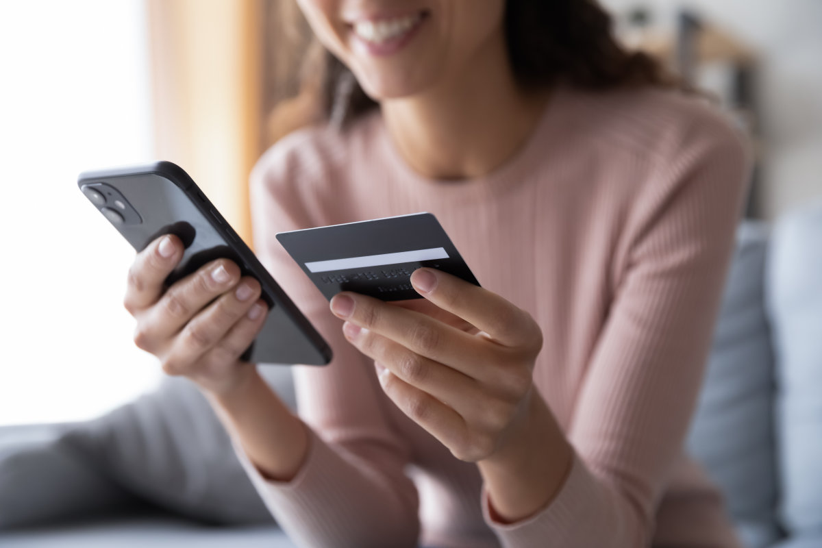 A woman smiling while holding a black smartphone in one hand and a black debit or credit card in the other.