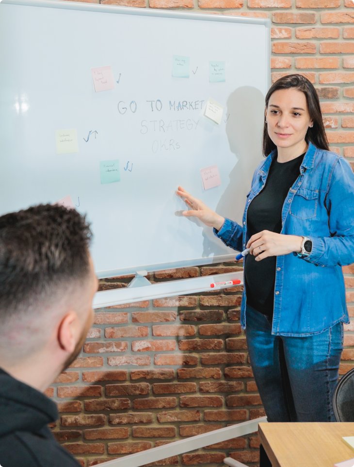 A woman in a denim shirt stands before a whiteboard with sticky notes and the words 'Go to Market Strategy OKRs' written on it. She gestures while speaking to a seated colleague. The setting is SumUp’s modern office in Sofia, Bulgaria