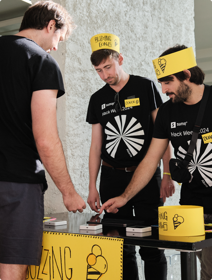 Two SumUp team members wearing Hack Week 2024 t-shirts and yellow paper hats labeled 'Buzzing Zone' stand behind a table with small devices, explaining their project to another participant during the event.
