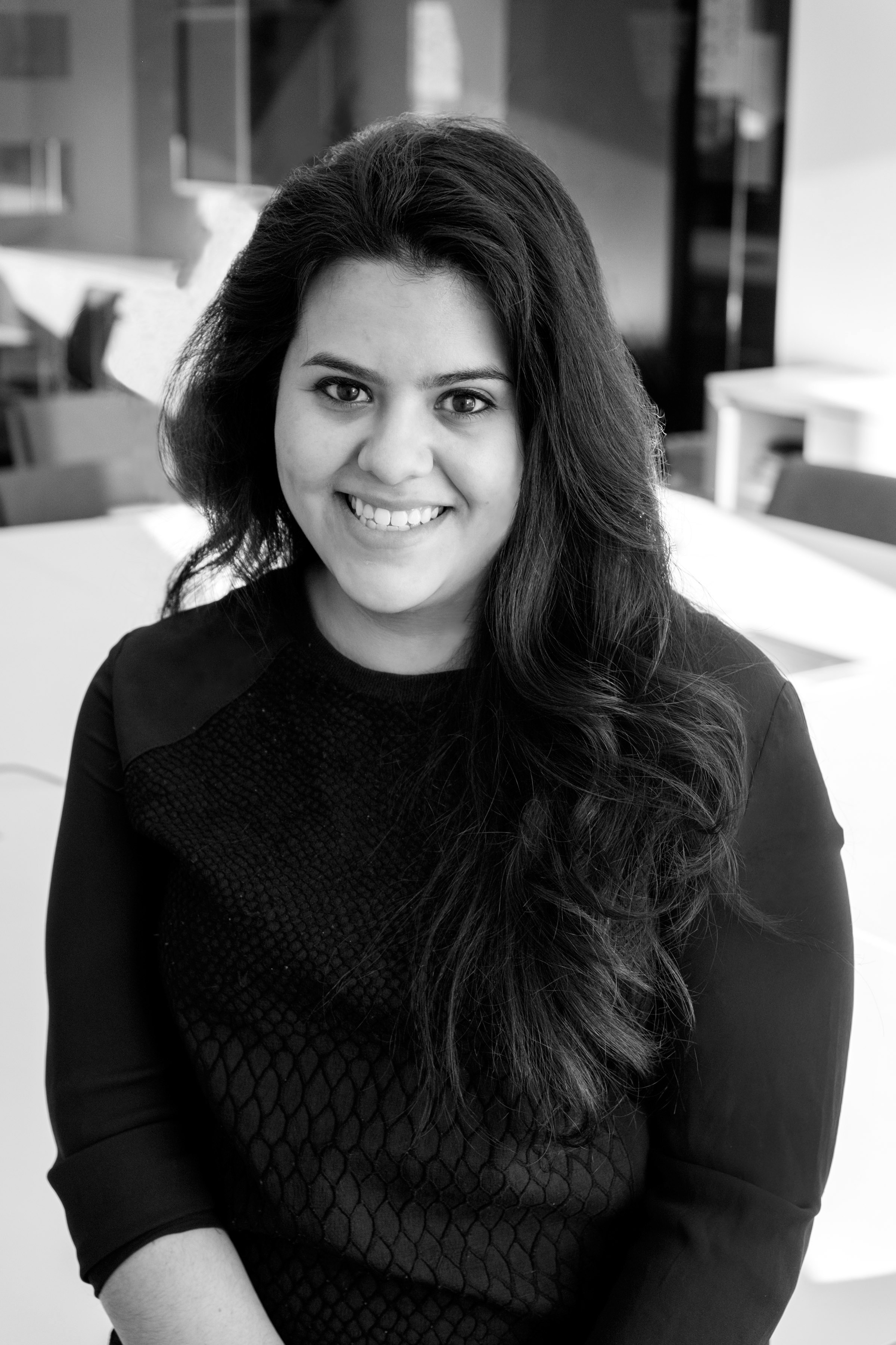 A person with long hair sits and smiles at the camera, wearing a dark top. The background is an indoor setting with blurred furniture and soft lighting.