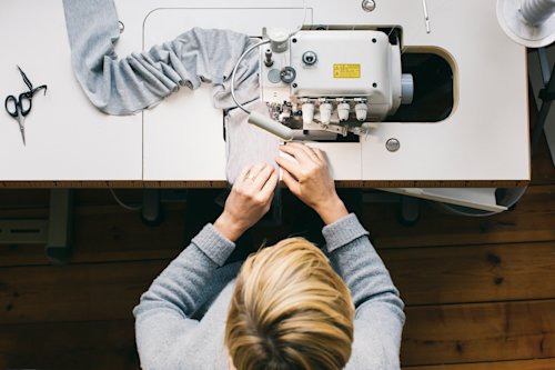 Hands sewing fabric using an industrial machine, surrounded by tools on a wooden table.