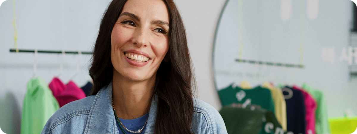 Femme souriante avec de longs cheveux noirs et qui porte une veste en jean, debout près d'une boutique de vêtements avec des tenues colorées en arrière-plan.