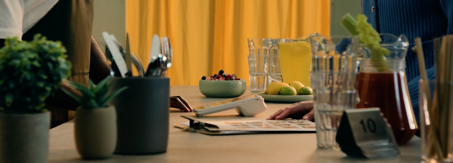 A SumUp handheld terminal sits on a counter at a restaurant among bowls of fresh fruit, glassware, and silverware.