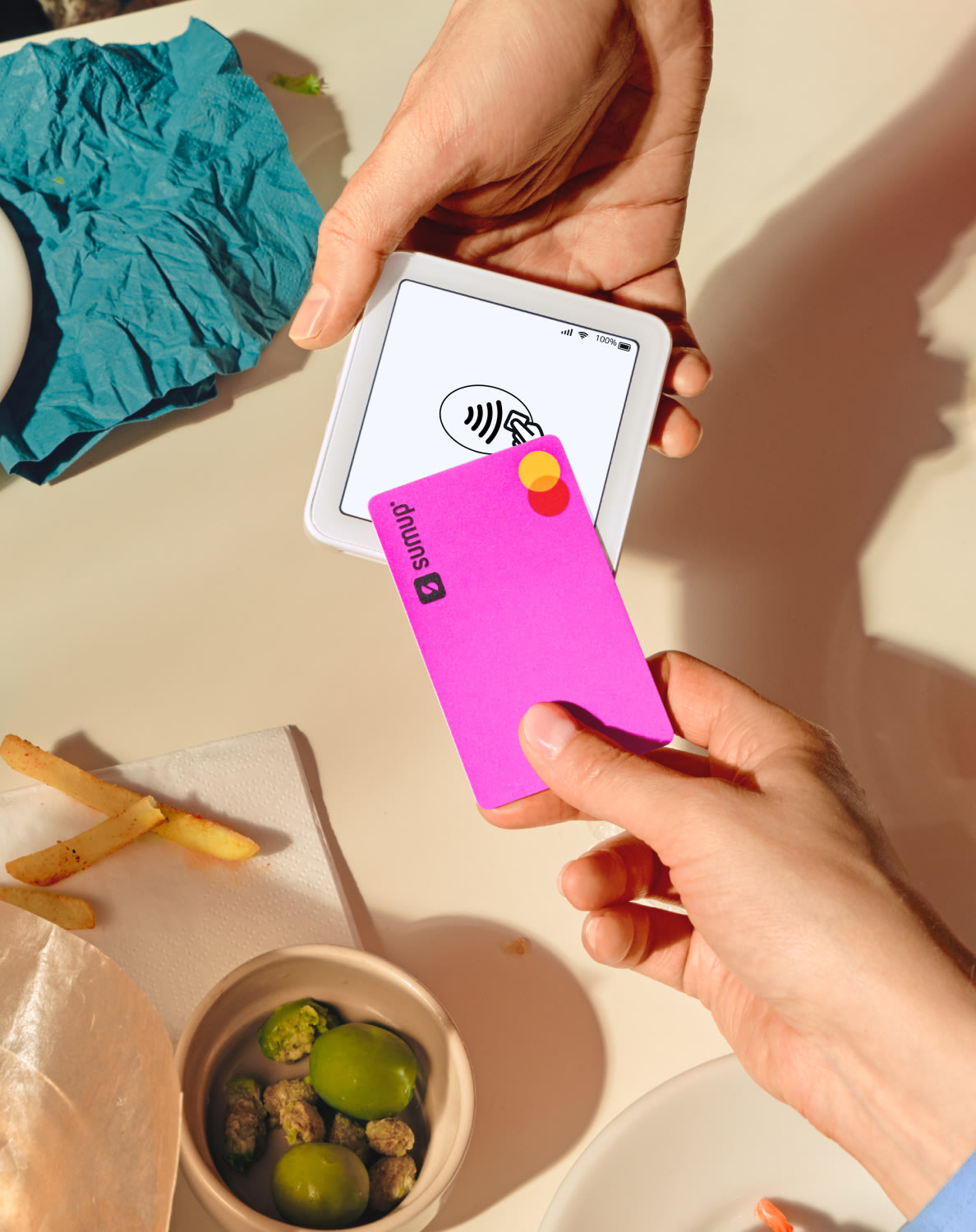 A hand holds a pink card near a SumUp Solo card reader, with food on the table and vibrant paper in the background