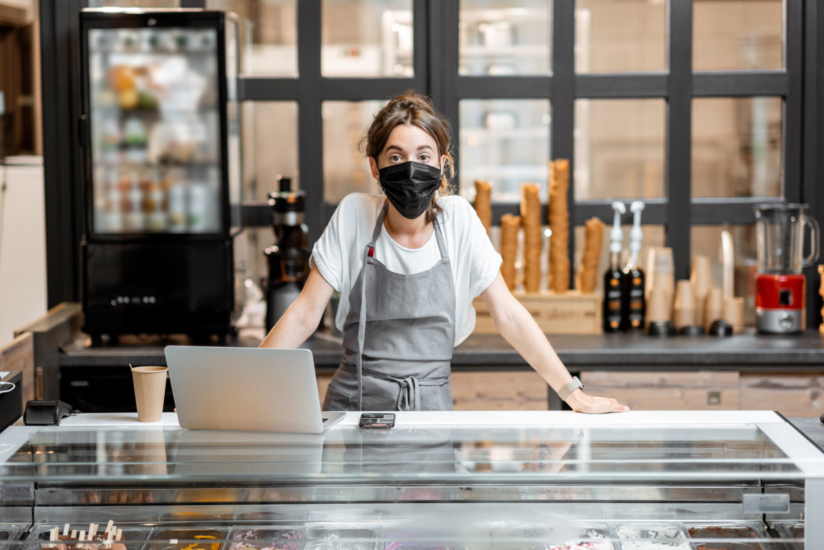 A woman wearing a protective mask stands behind the counter in an ice cream parlour. 